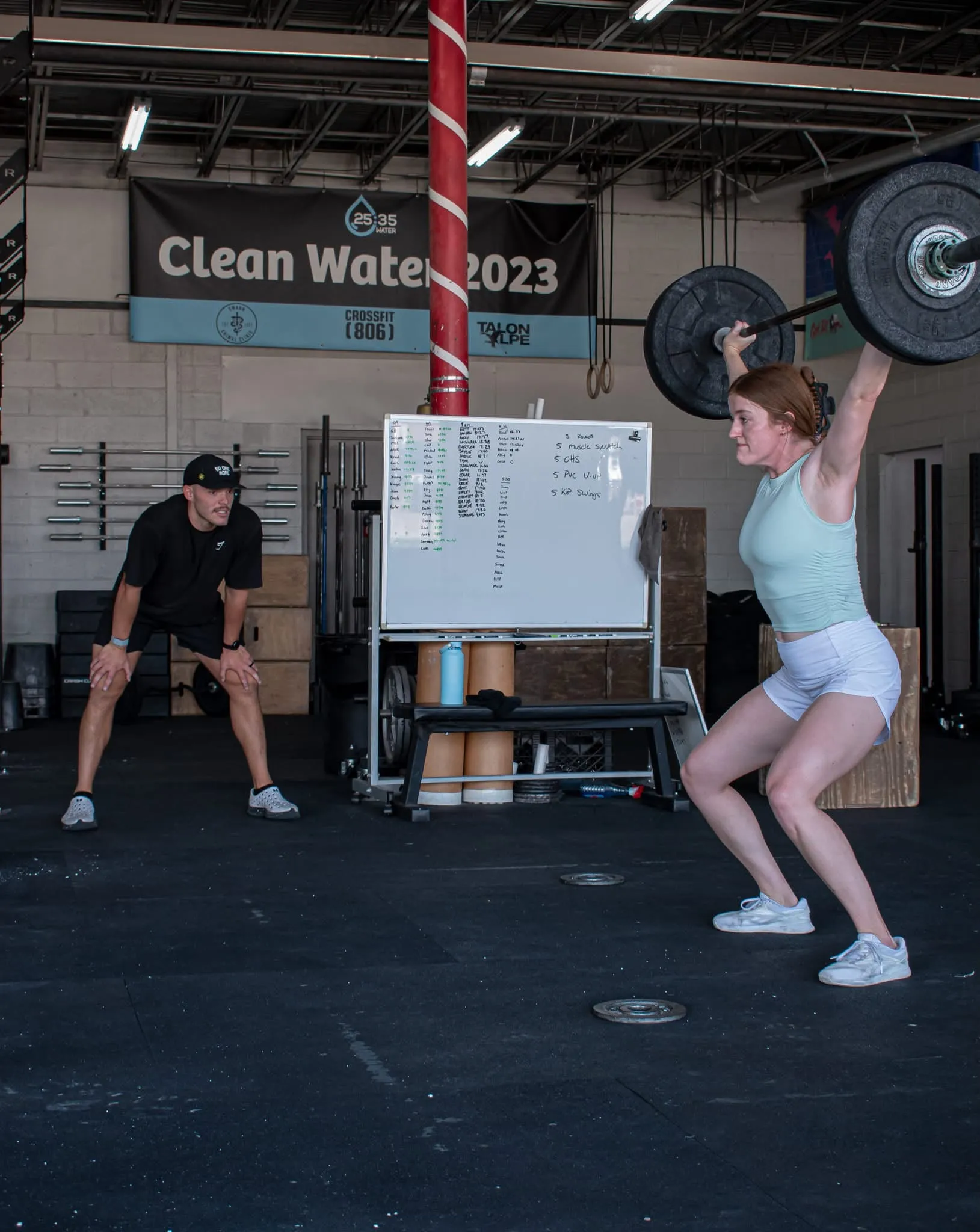 An athlete holding a barbell overhead while a coach watches her form at CrossFit 806